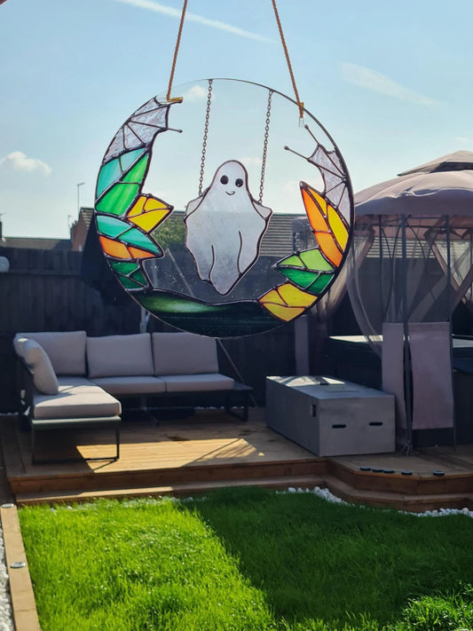Gothic stained glass sun catcher featuring an adorable ghost swinging within a crescent moon adorned with bat wings and vibrant autumnal leaves. Perfect for adding a spooky yet charming touch to any window or garden decor.