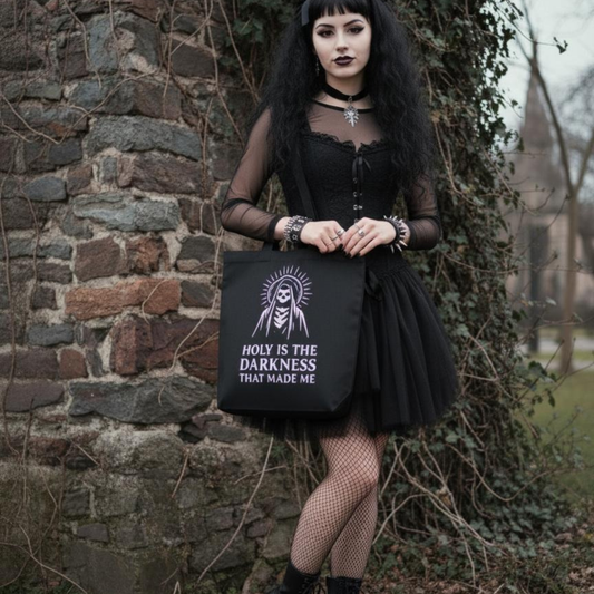 Person in gothic attire holding a black tote bag with text against a stone wall.