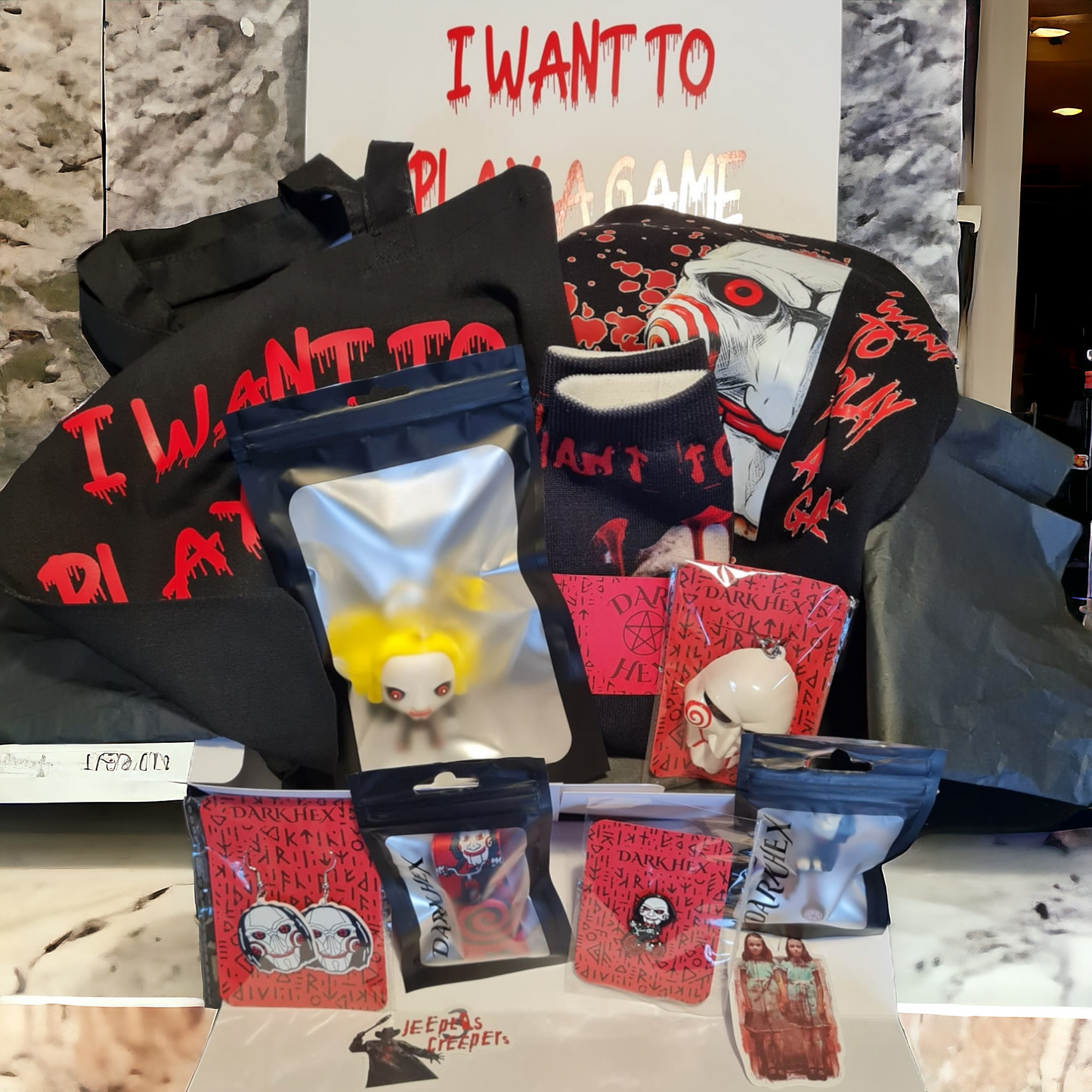 A variety of horror-themed merchandise including a black T-shirt with red text, a tote bag, socks with character designs, a keyring, a pin badge, a lanyard, earrings, a candle, and a fake blood-splattered gift note.